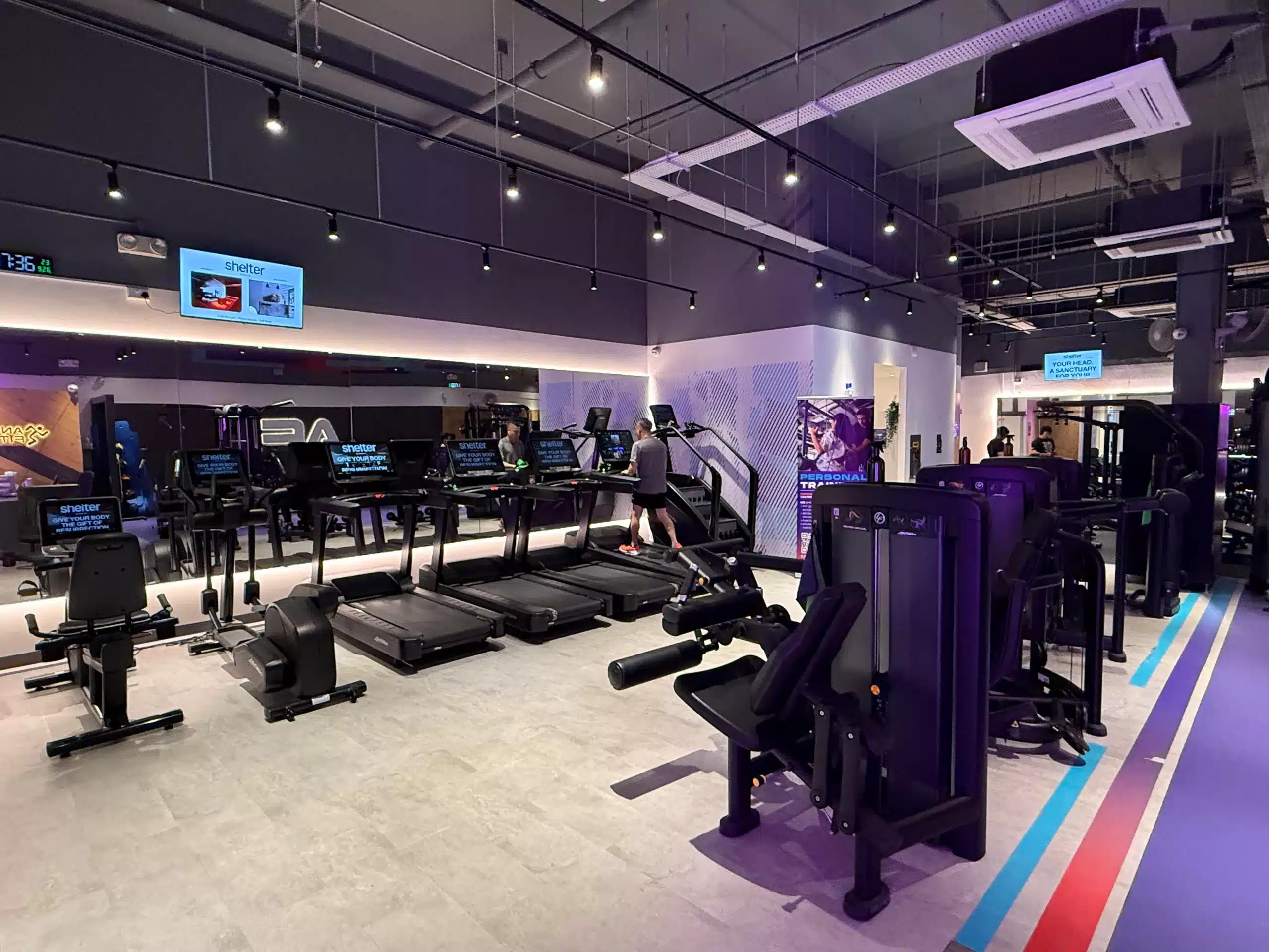 This image depicts the cardio area of Anytime Fitness Yishun East. The images on the screens of the treadmill and cardio machines have the words "Shelter by ReFormd". A row of leg machines are located behind the treadmills.