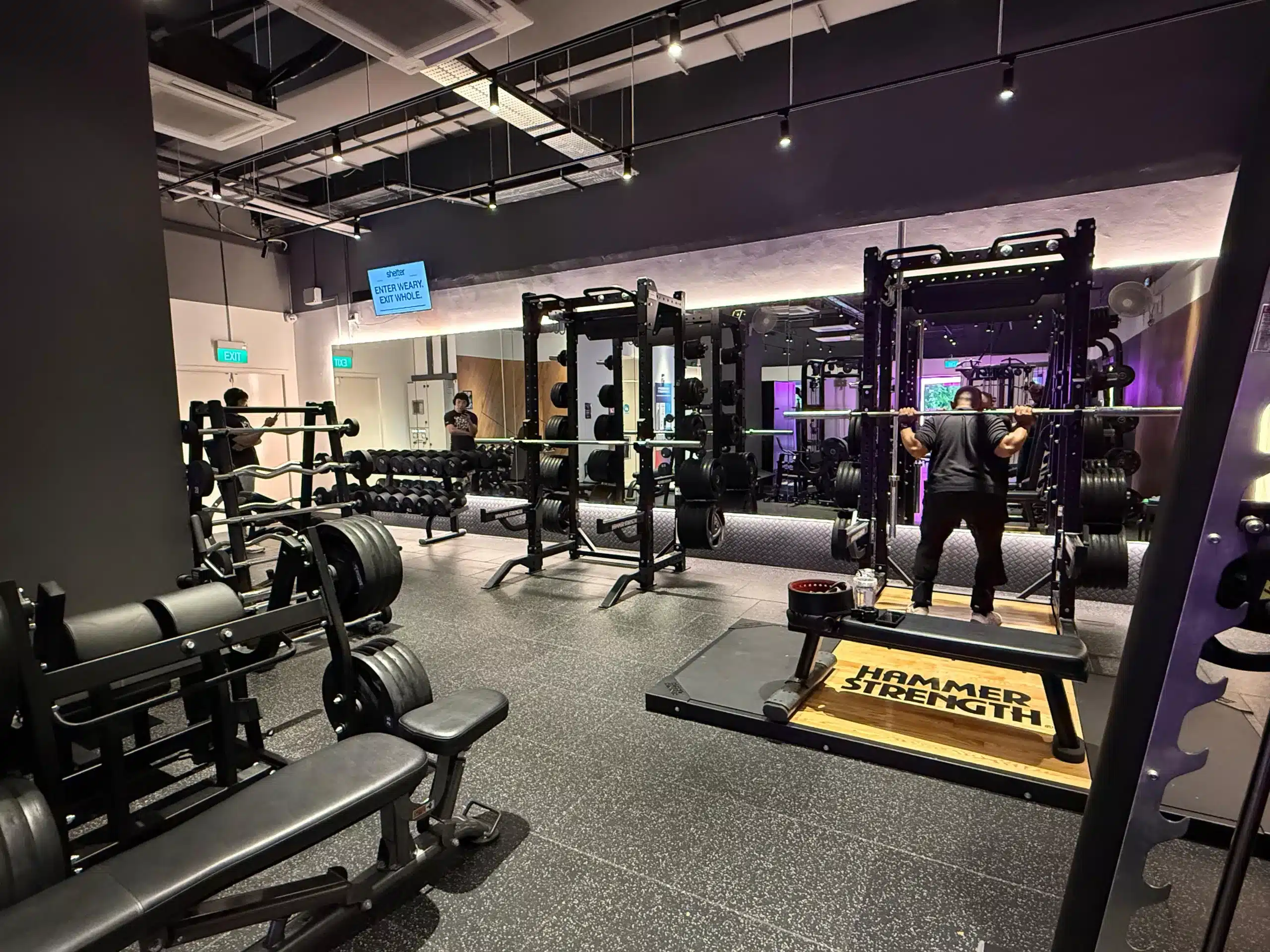 The free weight area of Anytime Fitness Yishun East. It depicts 2 racks with 1 rack having a member squatting with an empty bar. The rack that is being used has a deadlift platform in front of it.