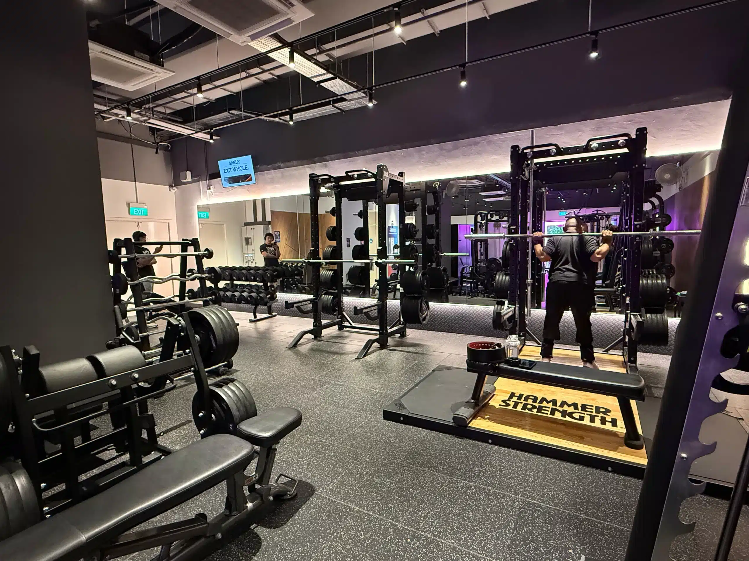 The free weight area of Anytime Fitness Yishun East. It depicts 2 racks with 1 rack having a member squatting with an empty bar. The rack that is being used has a deadlift platform in front of it.