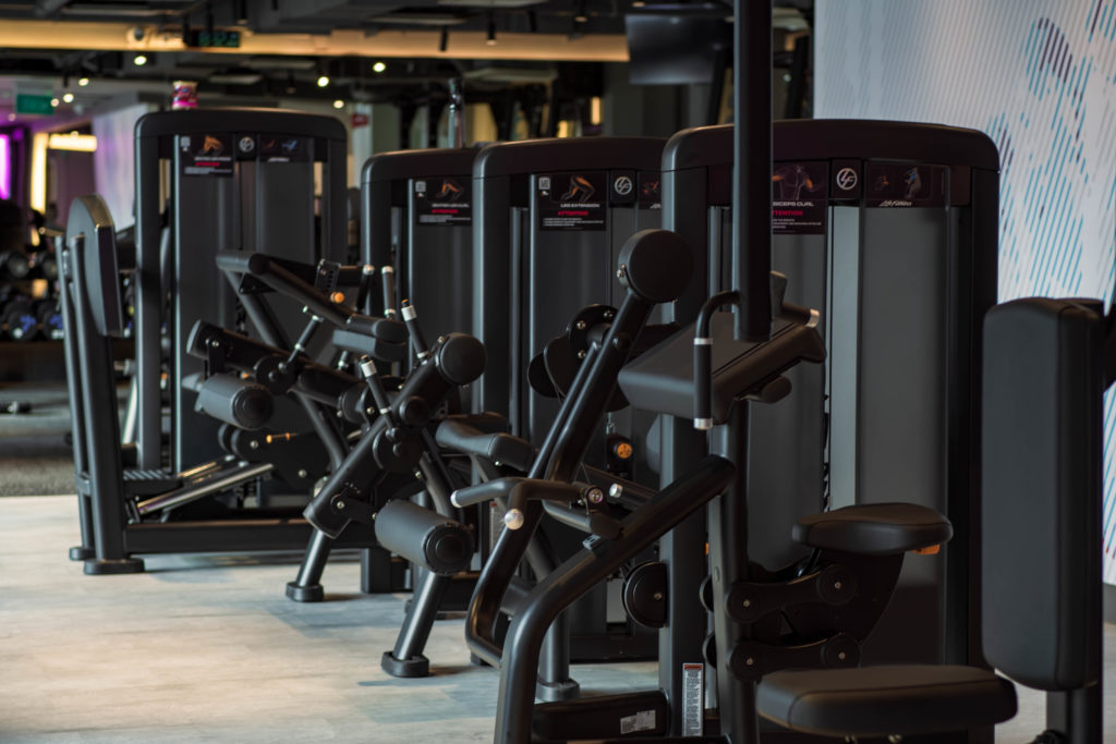 A row of modern gym machines, including a leg extension, preacher curl, pec dec, leg curl and leg press set in a bright, spacious fitness studio.