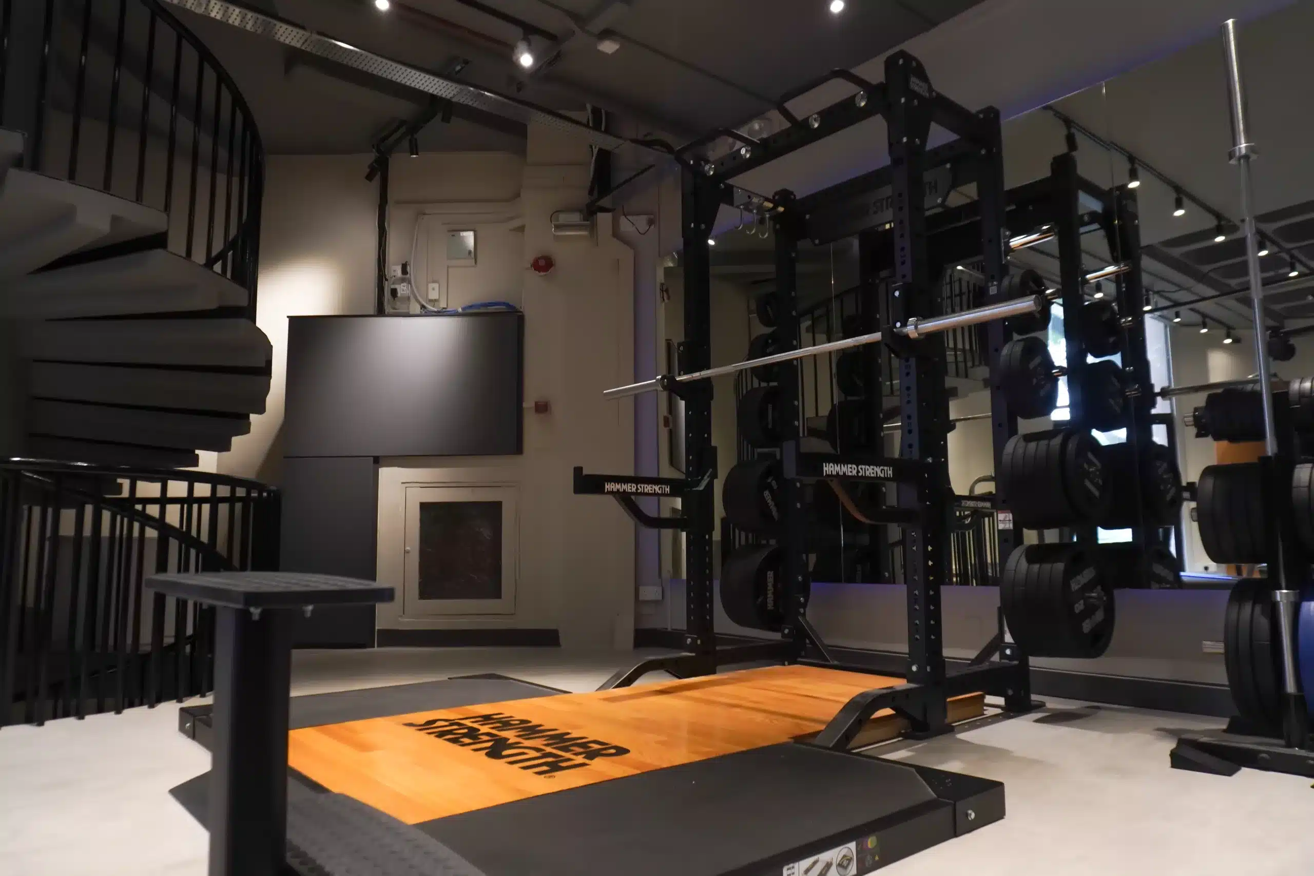 A lifting half-rack and deadlift platform located on level 1 of ReFormd Sixth Avenue. A barbell is placed in the rack and weight holders are full of hammer strength weights. A weight tree holder is slightly out of the picture, also filled with weights