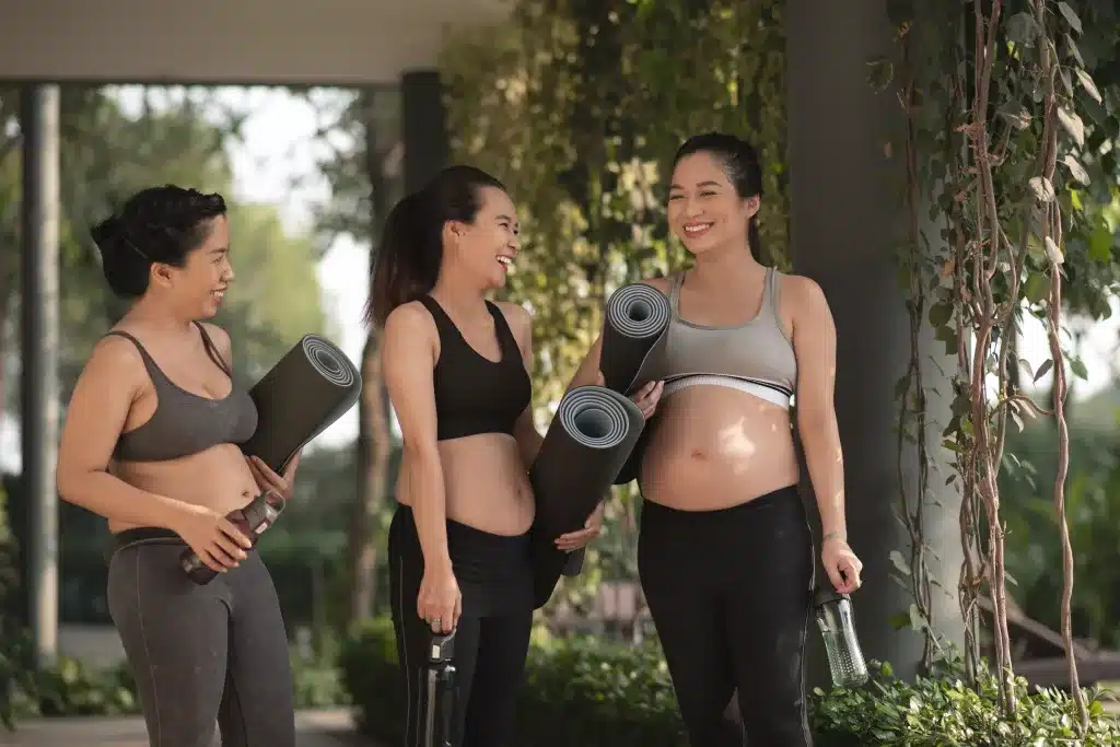 A group of 3 pregnant women laughing and holding yoga mats and water bottles.