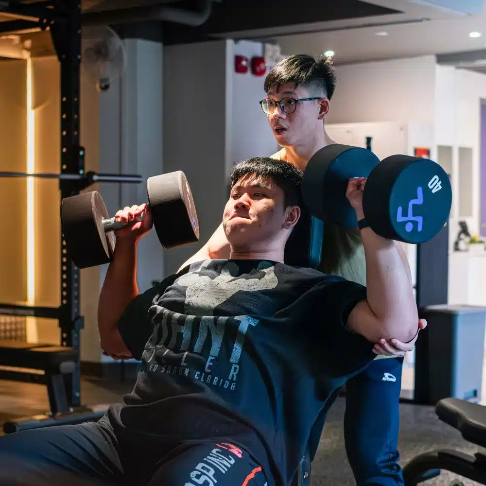 A well-built man executing a shoulder press with 40kg dumbbells. He is being supported by another man standing behind him with his hands placed below his elbows to help during the exercise